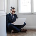 Ein Mann im Anzug sitzt auf dem Boden mit Laptop auf dem Schoß und lächelt