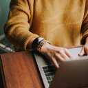 Frau in einem gelben Pullover sitzt vor ihrem Laptop und tippt auf der Tastatur